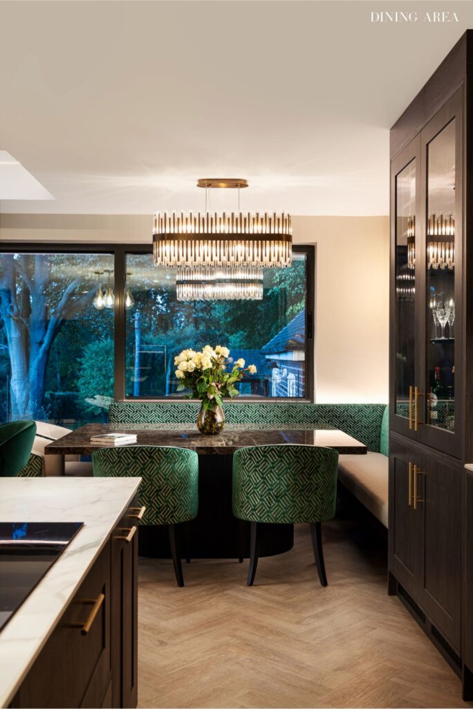 Elegant dining extension with large windows framing evening garden views, brass pendant lighting, green patterned dining chairs, dark cabinetry, and natural materials creating a sophisticated wellness-focused entertaining space by Katie Malik Interior Designer Essex Cambridge