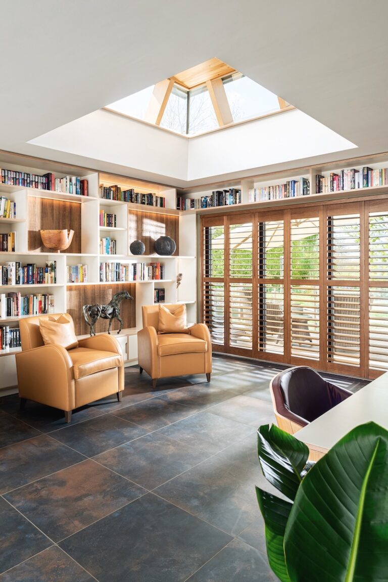 Bright library extension room with large skylight, built-in bookshelves, tan leather armchairs, wooden shutters, dark slate flooring, and potted green plants creating a wellness sanctuary with natural light and biophilic elements by Katie Malik Interior Designer Essex Cambridge