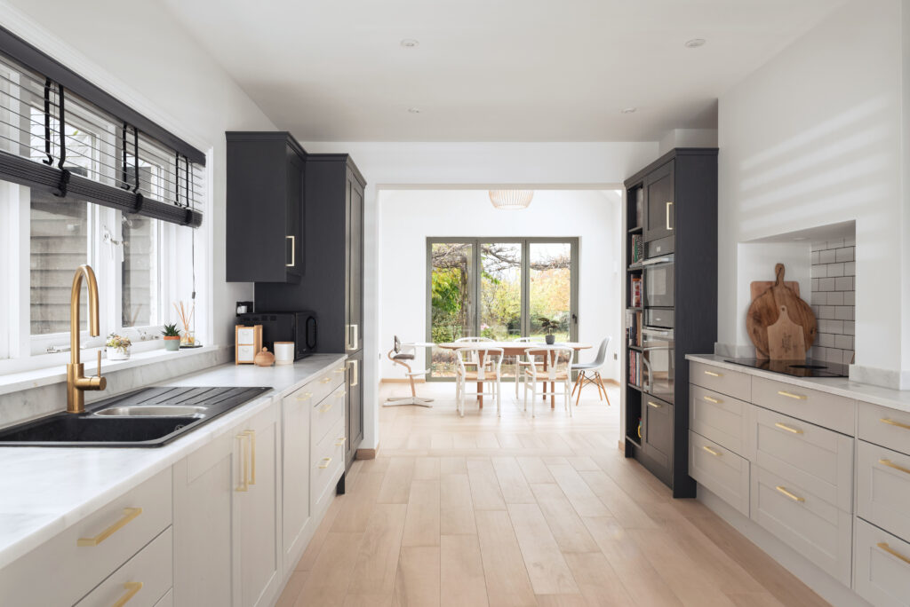 Minimalist contemporary kitchen extension with cream and black cabinetry, gold fixtures, large windows overlooking garden, and open-plan dining area with wooden chairs and natural light flooding through by Katie Malik Design Studio.