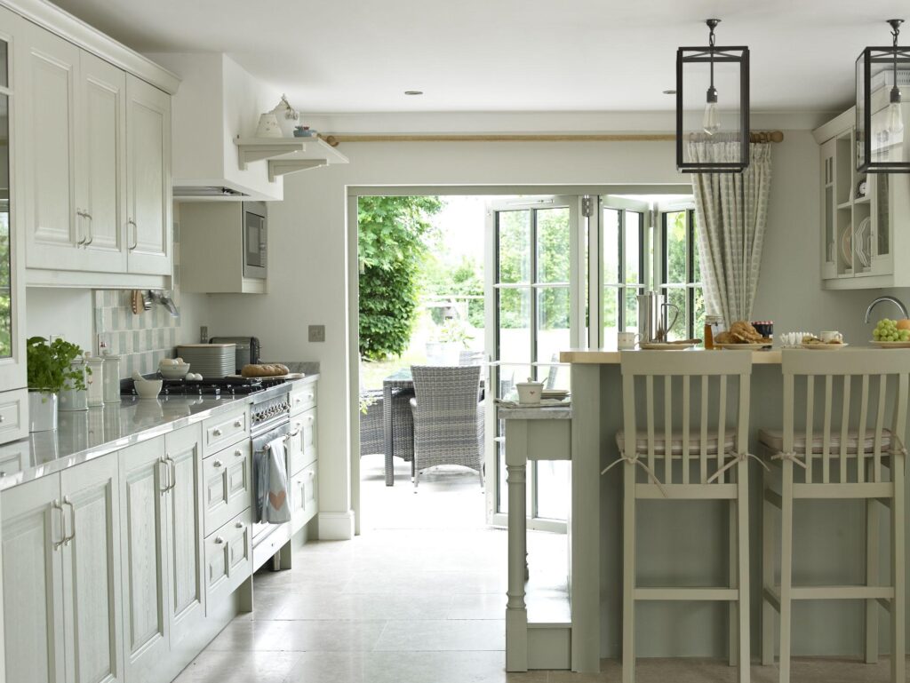 Bright modern kitchen extension with cream cabinetry, large bi-fold doors opening onto a garden patio, abundant natural light, and potted plants creating a biophilic connection to nature.