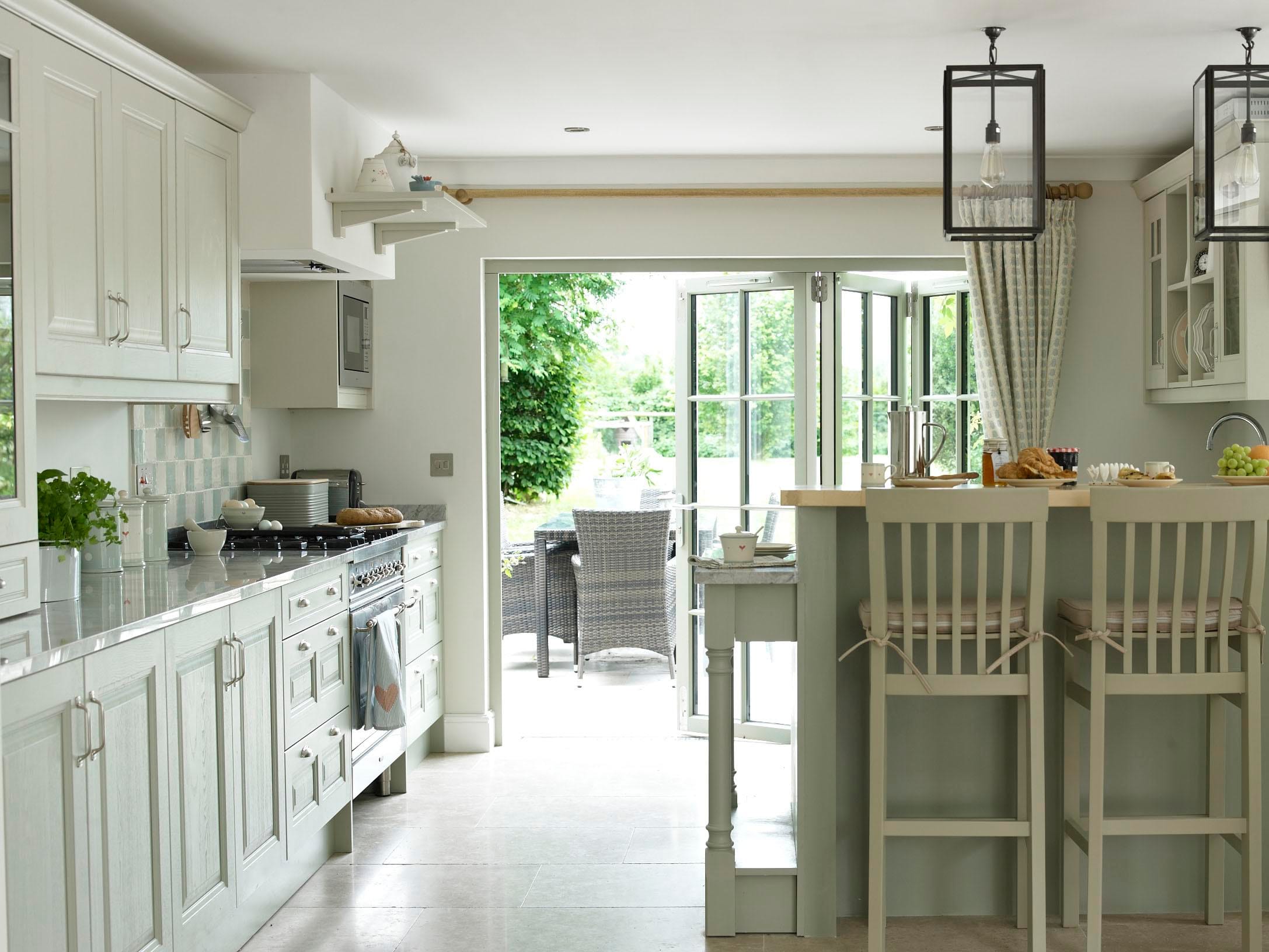 Bright modern kitchen extension with cream cabinetry, large bi-fold doors opening onto a garden patio, abundant natural light, and potted plants creating a biophilic connection to nature.