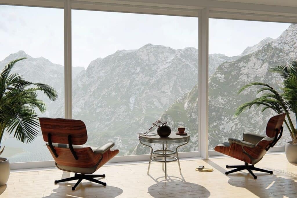 Two iconic Eames-style lounge chairs in walnut and leather flanking a chrome side table, positioned before floor-to-ceiling windows overlooking dramatic mountain landscape, with palm plants on either side