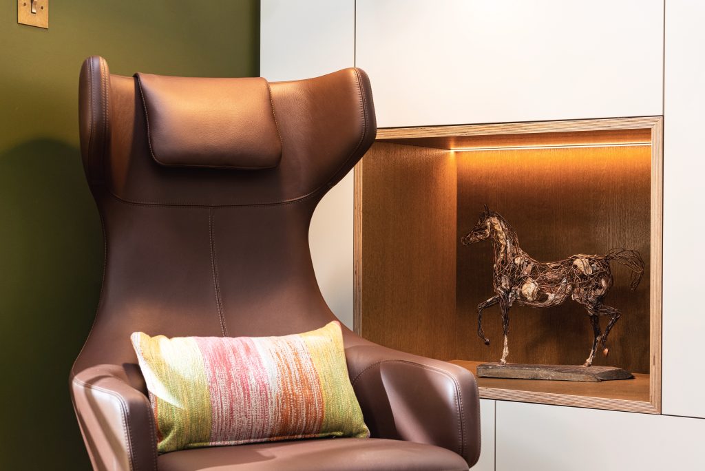Luxury brown leather wingback chair with headrest pillow, colorful striped cushion, and wooden framed niche displaying bronze horse sculpture with warm lighting- Interior designer Essex and Cambridge- Katie Malik Design Studio