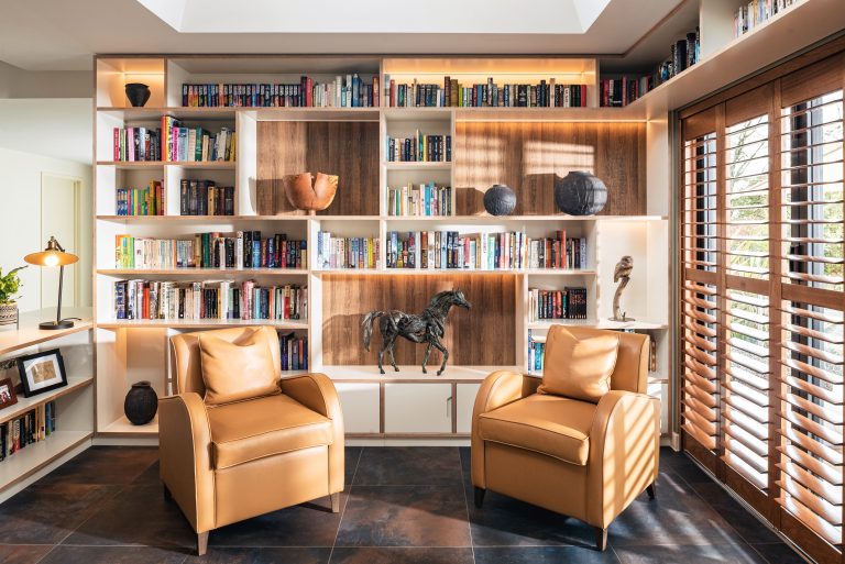 Contemporary library with floor-to-ceiling white shelving, warm wood backing, two tan leather armchairs, bronze horse sculpture, warm accent lighting, and wooden shutters- Interior Designer Essex- Katie Malik Design Studio