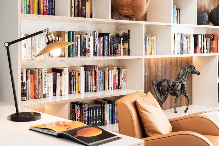 Modern workspace with white shelving, colorful books, bronze horse sculpture, tan leather chair, brass desk lamp, and open books on white desk- Essex Interior designer