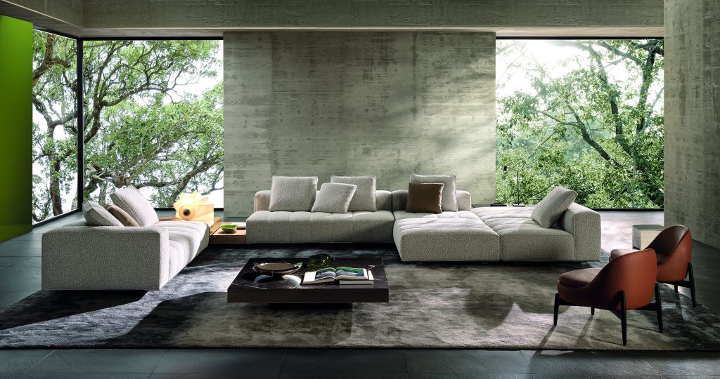 Contemporary luxury living room designed by Essex interior designer Katie Malik, featuring bronze upholstered sofas, dark burgundy accent wall, floor-to-ceiling windows with mountain views, concrete ceiling beams, and sophisticated neutral furnishings creating a serene sanctuary.