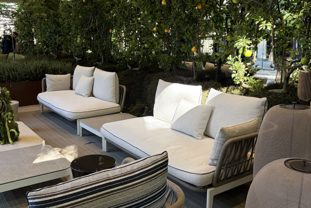 A biophilic interior design setting featuring white modular outdoor sofas with woven rope detailing, a striped armchair, and mesh floor lamps, all surrounded by lush indoor trees and natural light.